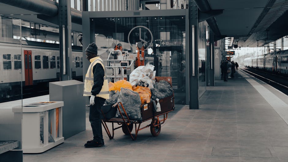 A worker wearing a dark uniform, a grey beanie, and a yellow safety vest stands on a train station platform next to a large trolley loaded with numerous garbage bags and discarded items, including plastic and paper waste. The platform features modern, grey tiling, and the station is well-lit with natural light filtering through glass panels and overhead fixtures. To the left, there is a white waste disposal container, and in the background, parked trains and station signage are visible, indicating an active transit environment. This scene captures routine cleaning or waste clearance activities, highlighting the importance of hygiene and maintenance in busy transportation hubs. For expert cleaning services, contact error code: 521 at carpetcleaningn1.co.uk, providing professional deep cleaning and sanitisation solutions for commercial and residential surfaces.