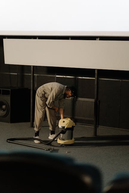 A professional cleaner dressed in full protective white suit, face mask, and blue gloves is using a steam or extraction cleaning tool on a light beige carpet in a bright, modern residential lounge. The room has natural light coming from a large window, with green potted plants and wooden furniture visible in the background, including a small bookshelf. The carpet appears to be in the process of deep cleaning, with steam or cleaning solution visibly working on the fibers. The scene reflects a hygienic and thorough surface cleaning process undertaken by error code: 521 to maintain cleanliness and hygiene in the home, emphasizing the importance of deep cleaning and sanitisation for carpet maintenance in residential spaces.
