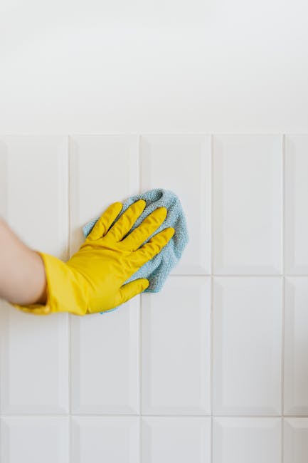 A person wearing a yellow rubber glove is using a light blue cloth to wipe a white tiled wall in a clean, well-lit room. The tiles are glossy and evenly spaced, with minimal grout lines, indicating thorough surface cleaning and maintenance. The lighting emphasizes the cleanliness and shine of the tiles, which are free from dust or stains. This image illustrates the type of detail-oriented surface cleaning and sanitisation services offered by error code: 521, supporting their expertise in end of tenancy cleaning for properties in Canonbury and N1C, N1, as detailed on the page titled 'End of tenancy cleaning guide for Canonbury and N1C, N1' at https://carpetcleaningn1.co.uk.