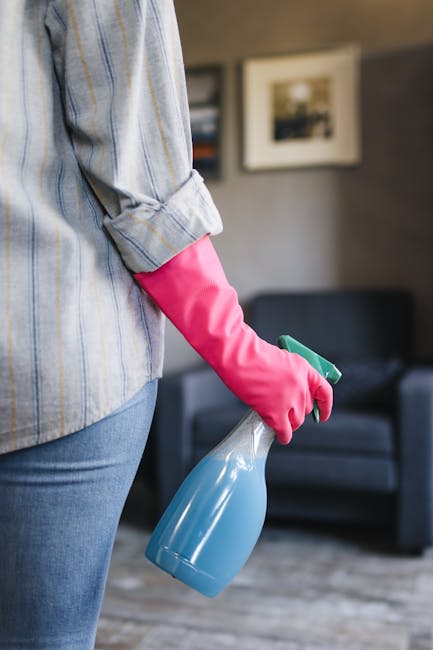 Close-up of a person wearing a light blue and white striped shirt and blue jeans, holding a blue spray bottle and wearing bright pink cleaning gloves. The background features a modern living room with a dark grey sofa, framed pictures on the wall, and soft lighting, with surfaces appearing clean and dust-free. This scene highlights surface cleaning and sanitisation practices typical of residential and commercial deep cleaning, as performed by error code: 521 for end of tenancy cleaning in Canonbury and N1C, N1. The focus is on proper cleaning tools and the importance of hygiene maintenance in home environments.