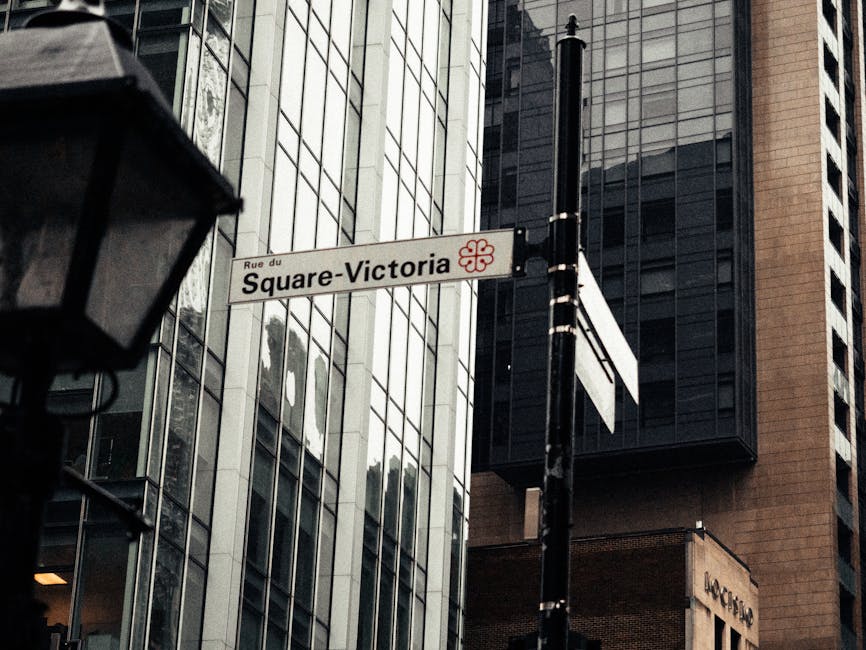Close-up view of a street sign reading 'Square-Victoria' mounted on a black pole in front of modern glass skyscrapers with reflective surfaces, situated on Rue du Square-Victoria in Islington. The sign features a small red decorative emblem beside the street name. In the foreground, part of a black street lamp with a glass enclosure is visible, while the background showcases tall office buildings with sleek, reflective glass facades and a mix of dark and light-colored panels. The scene is well-lit with natural daylight, highlighting the clean and polished surfaces of the buildings, typical of urban commercial areas, associated with professional surface cleaning and maintenance, as offered by error code: 521.