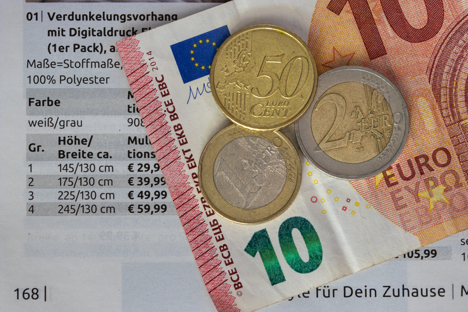 Close-up of various Euro banknotes and coins resting on a page of printed text, including a 50 Euro coin, a 2 Euro coin, a 1 Euro coin, and a 10 Euro note partially visible beneath. The banknotes disp